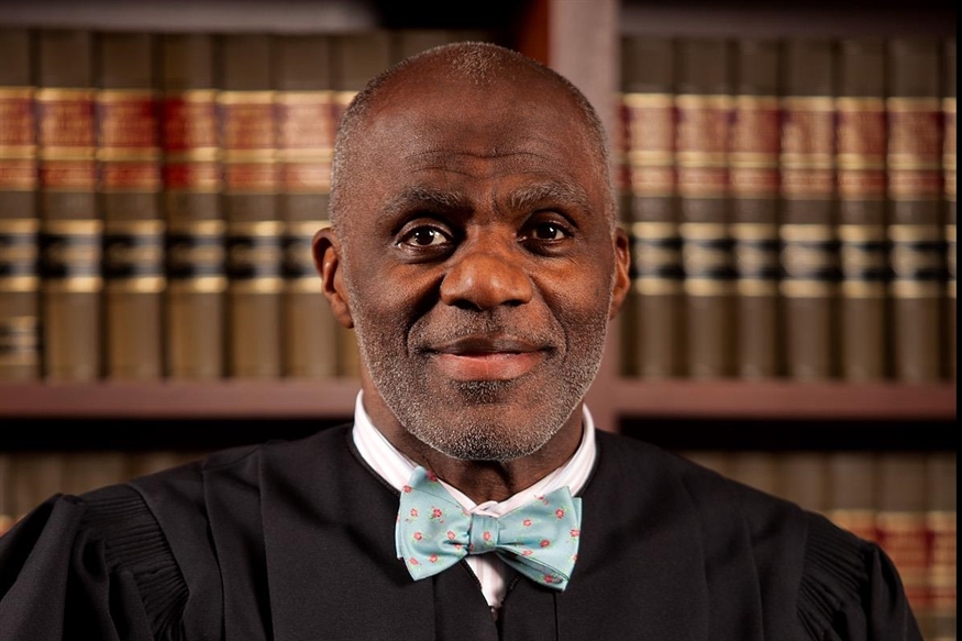 [ai] A smiling judge wearing a black robe and a light blue bow tie, standing in front of a large bookshelf filled with legal books.